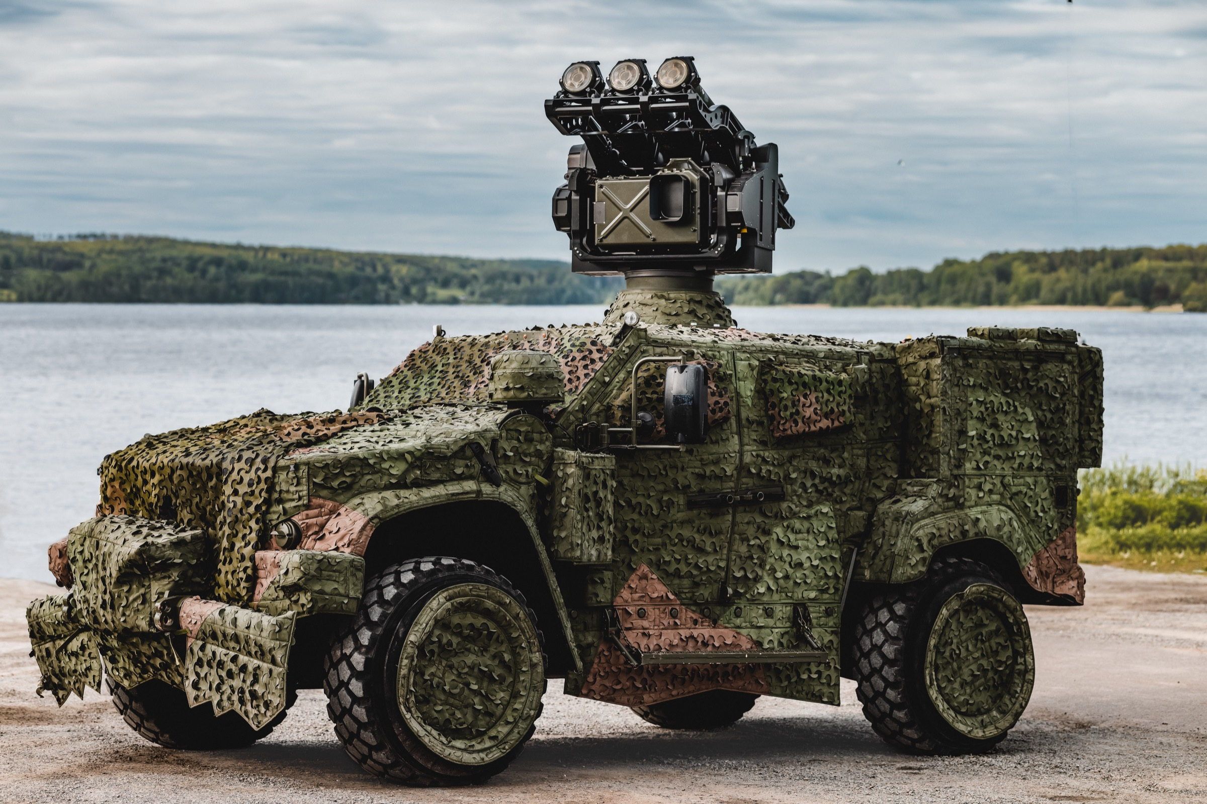 A camouflaged Oshkosh vehicle carrying a mast-mounted RBS 70 NG launcher and sight, parked by a lake